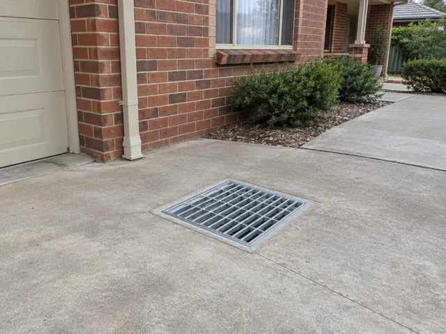 Outdoor driveway drain grate set in dry concrete beside a brick house in Weston